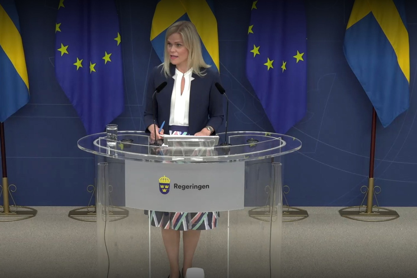 Person står vid ett glasbord som det står Regeringen på. Bakom syns två svenska och två EU-flaggor.