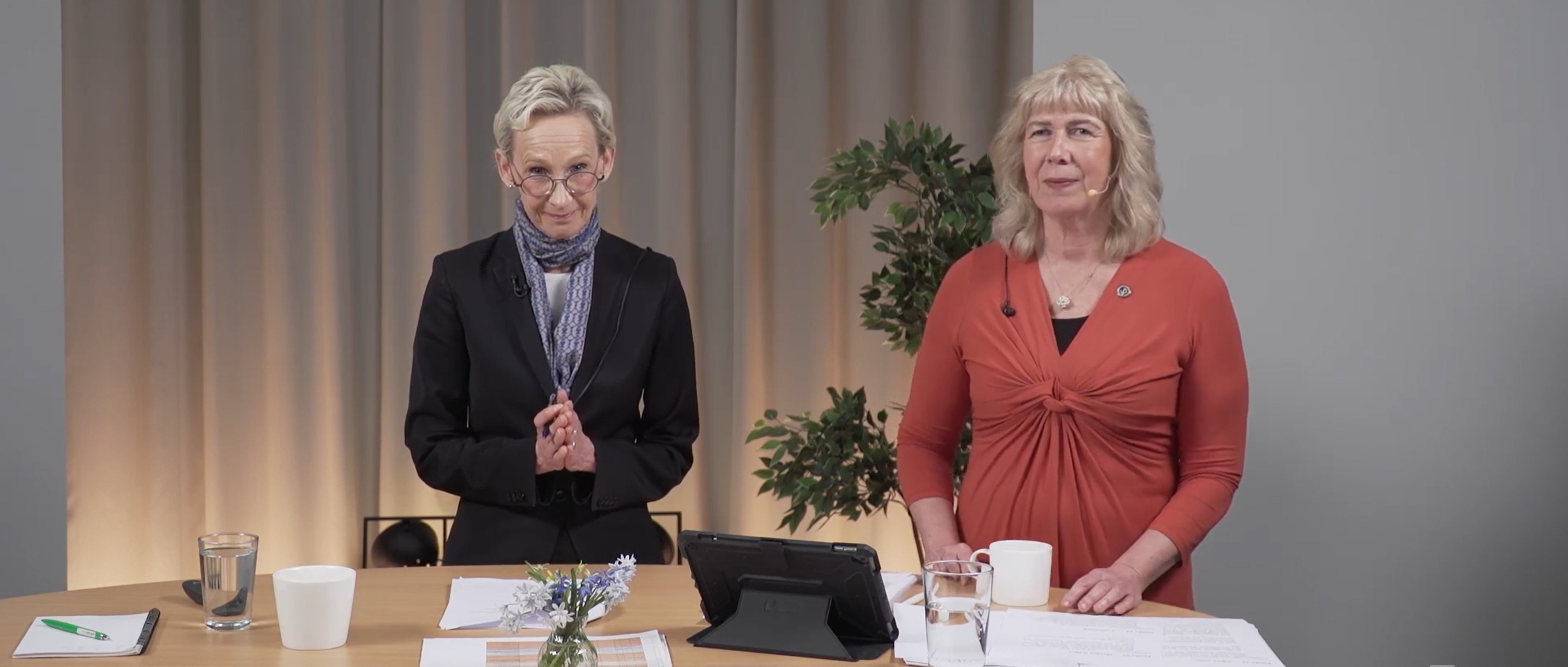 Två personer står i en studio. På bordet framför dem ligger papper och står tekoppar och vattenglas. Bakom syns en hög grön växt.