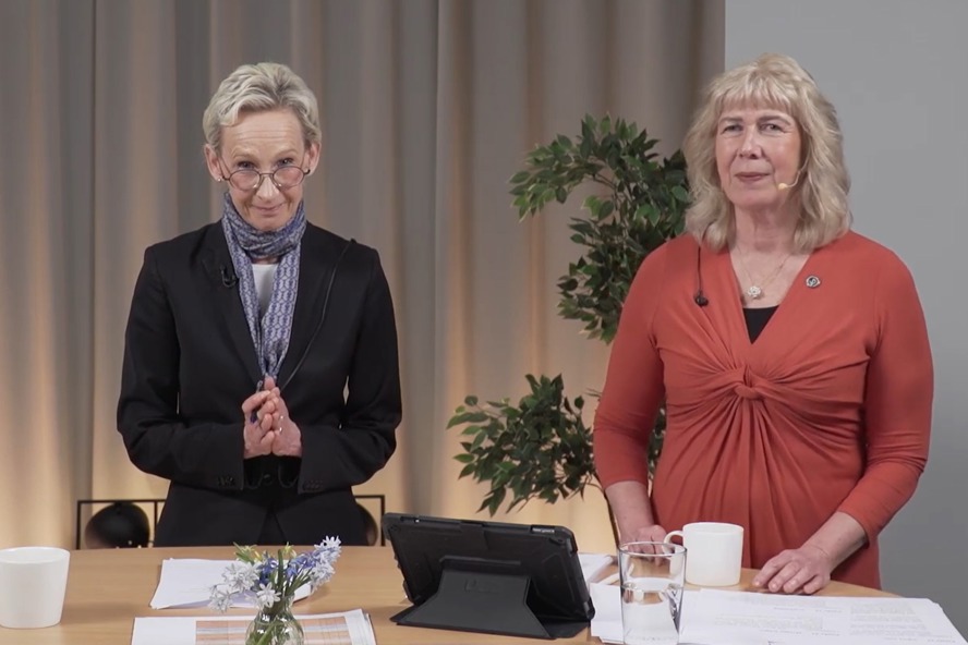 Två personer står i en studio. På bordet framför dem ligger papper och står tekoppar och vattenglas. Bakom syns en hög grön växt.