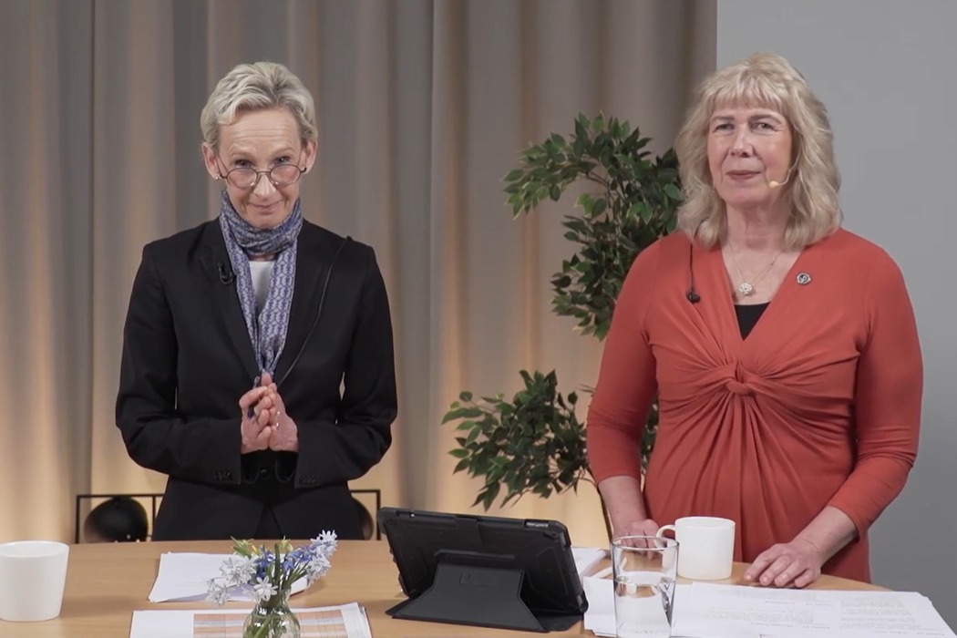 Två personer står i en studio. På bordet framför dem ligger papper och står tekoppar och vattenglas. Bakom syns en hög grön växt.