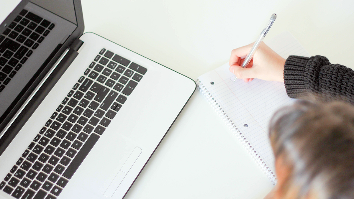 Person sitter framför dator och skriver samtidigt i ett anteckningsblock.