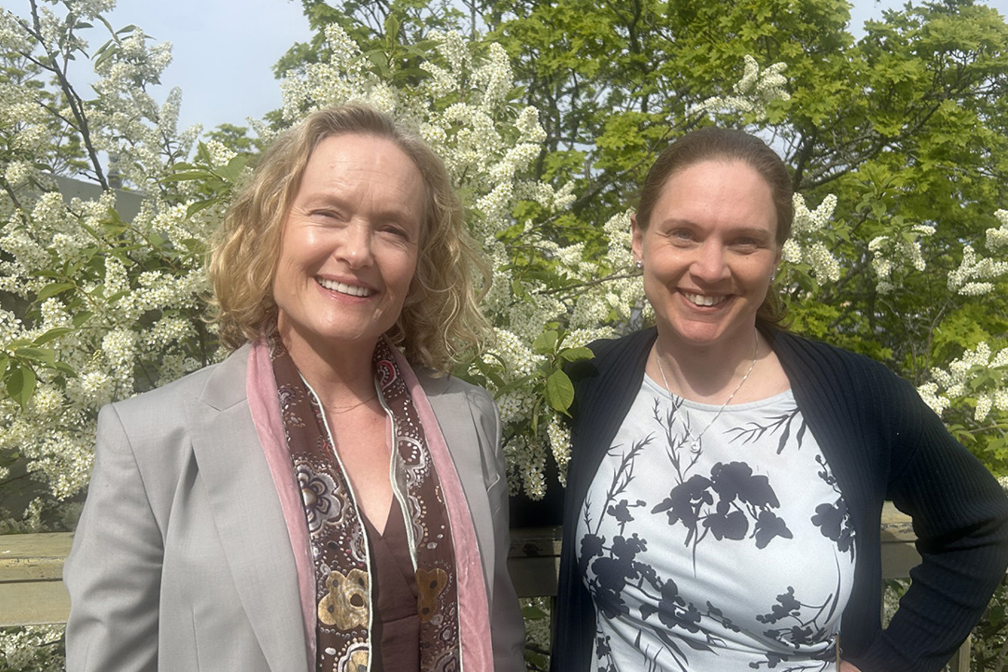 Två personer står utomhus framför en blommande hägg. De ler och tittar in i kameran.