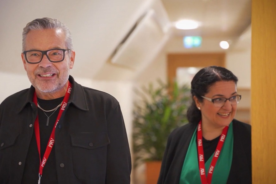 Två personer kommer gående i en korridor, de har nyckelband från PRV runt halsen. 