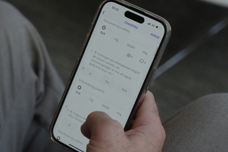 Person sitter med en mobil i handen. I mobilen syns en digital checklista från IA-systemet som kan fyllas i genom reglageknappar.