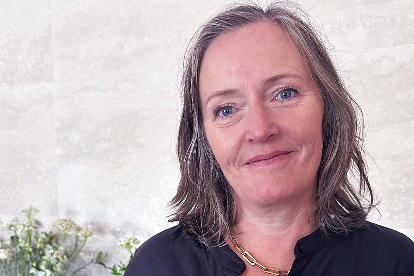 Person iförd svart blus och halsband, stående framför en bukett blommor.