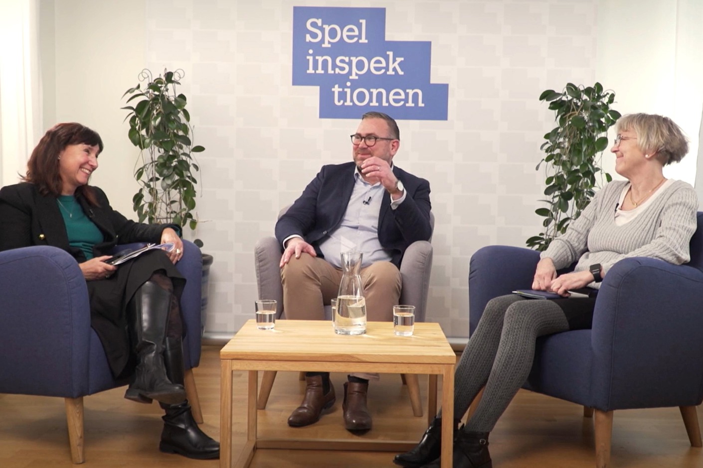 Tre personer sitter i fåtöljer i en studio. Bakom dem syns Spelinspektionens logotyp. De ler.