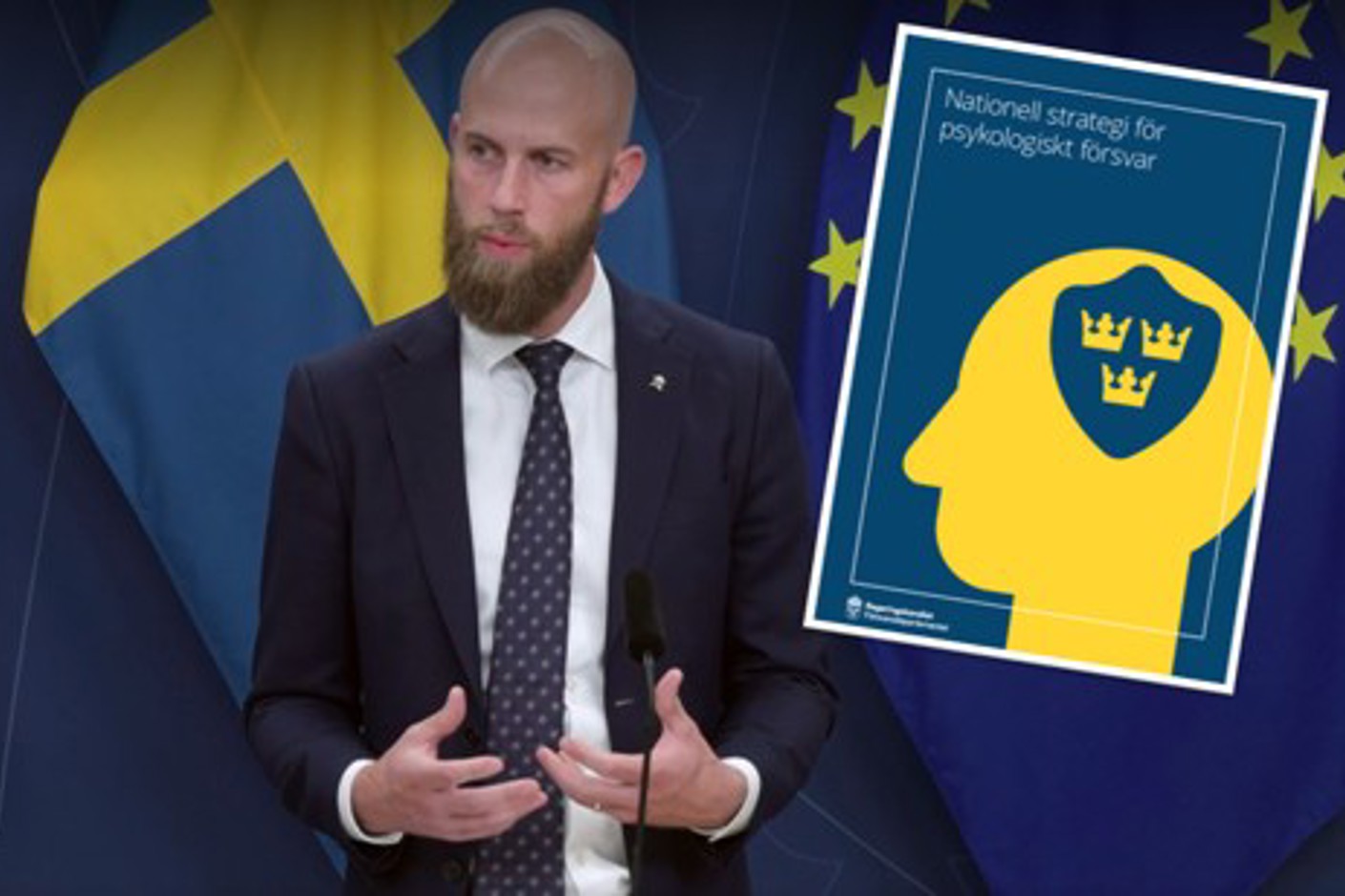 Ministern för civilt försvar Carl-Oskar Bohlin står framför svensk flagga och EU-flaggor. Till höger om honom syns omslaget på den nya nationella strategin för psykologiskt försvar