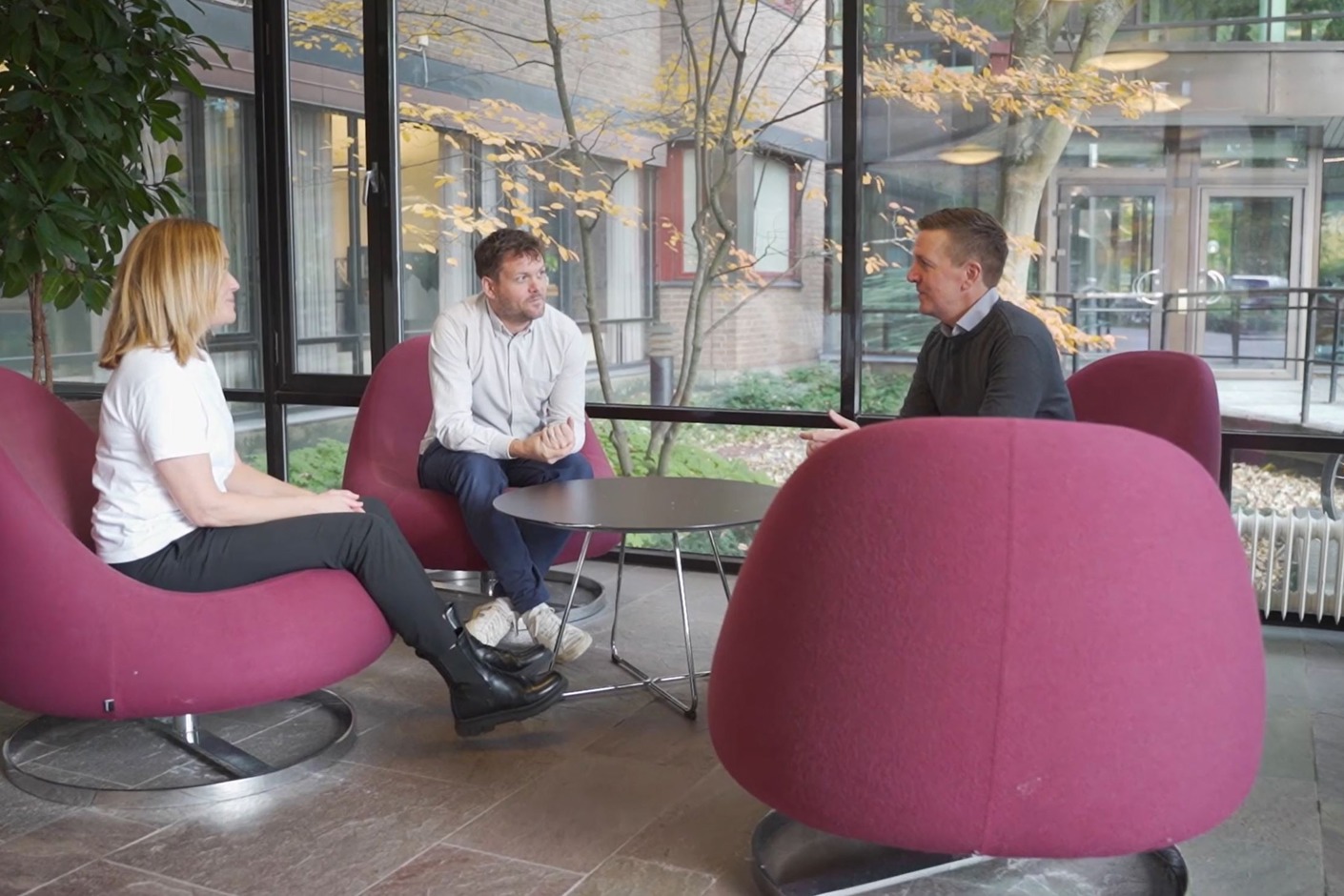 Tre personer sitter i fåtöljer i en glasad lobby. Bakom syns träd och cyklar.