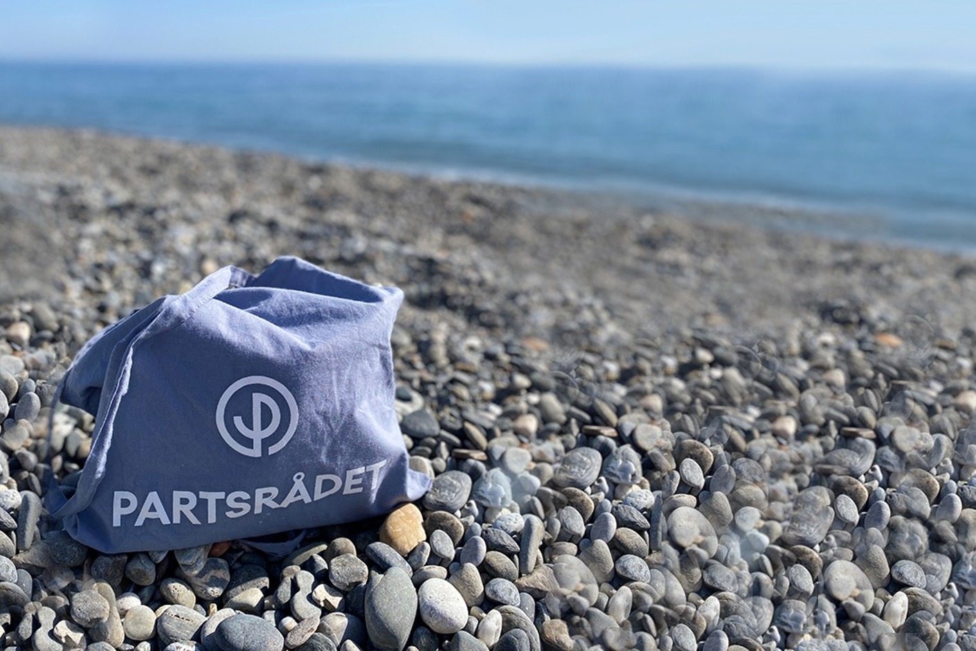 En blå tygpåse med Partsrådets logotyp som står på en stenstrand i Spanien.