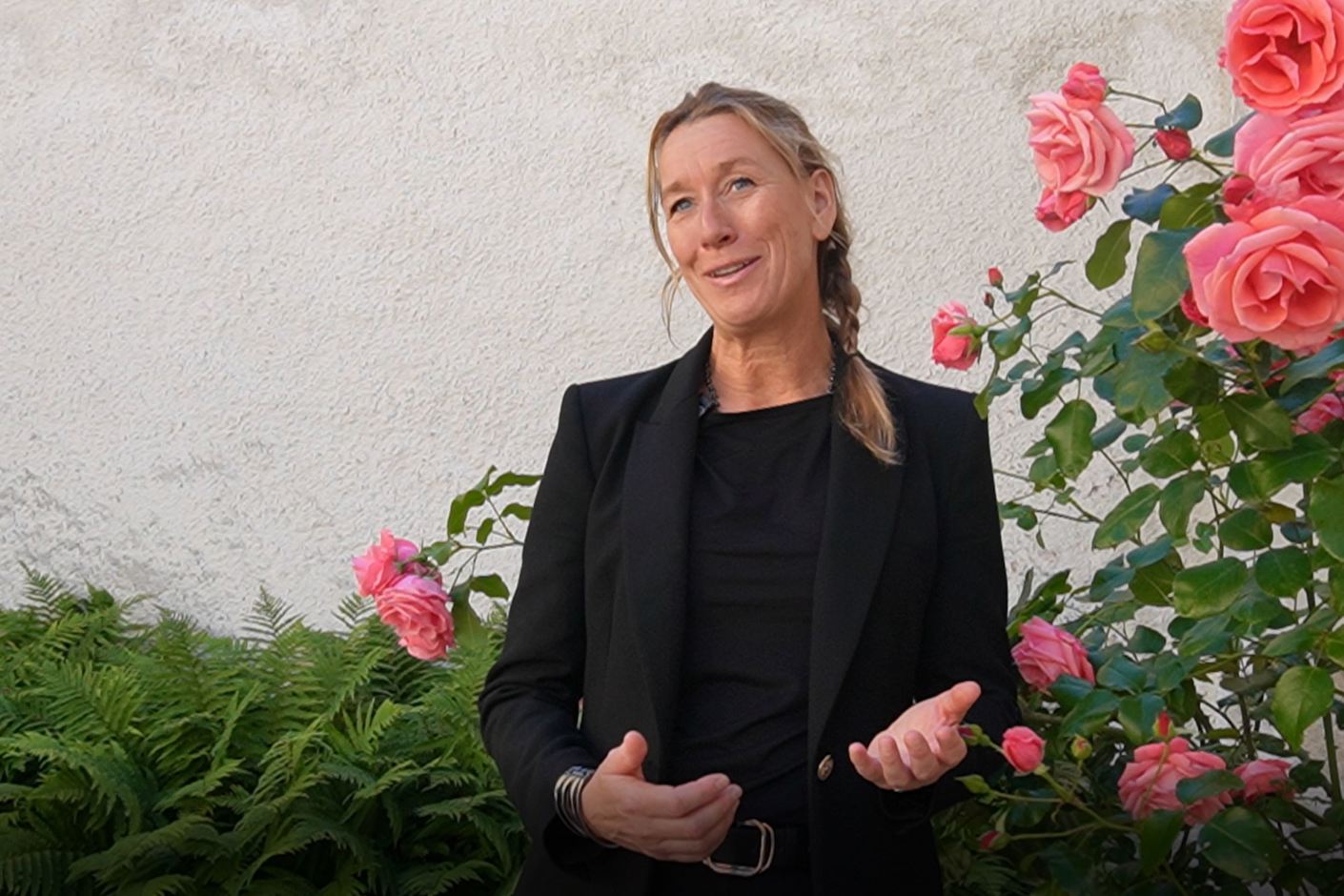 Kvinna står och pratar in i kameran utomhus med rosor bredvid sig.