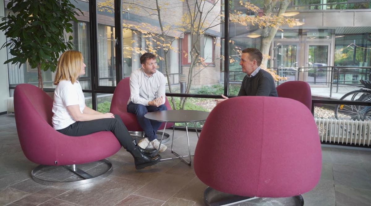Tre personer sitter i fåtöljer i en glasad lobby. Bakom syns träd och cyklar.