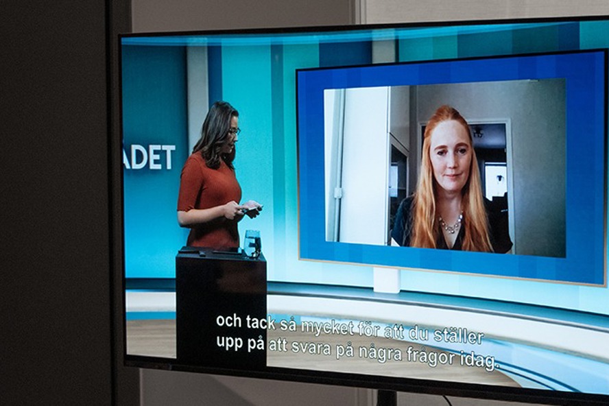 En person stående i studiomiljö intervjuar en person som deltar i sändningen via länk.
