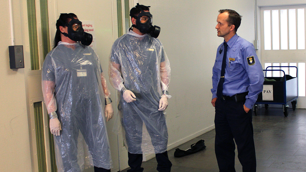 Två personer med skyddsutrustning (gasmask och plastöverdrag över kläderna) och en person klädd i Kriminalvårdens kläder.