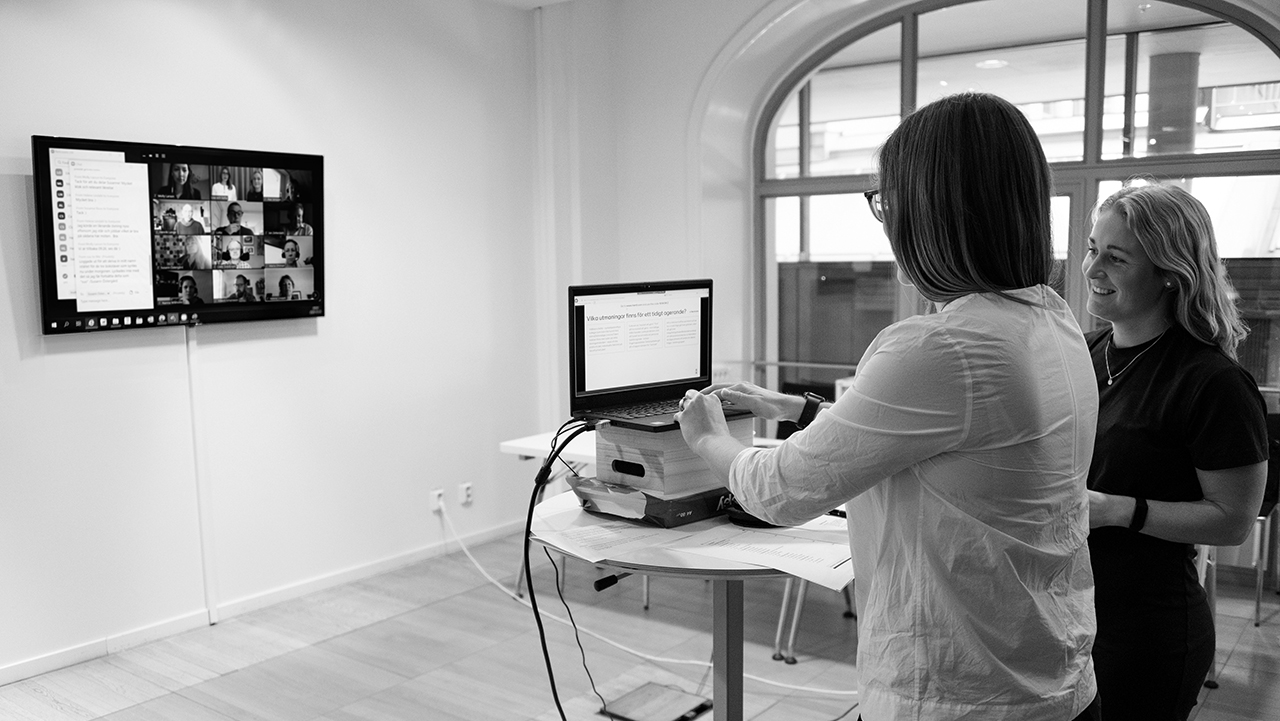 Två personer framför ett ståbord med ett Temasmöte pågående på en skärm på väggen.