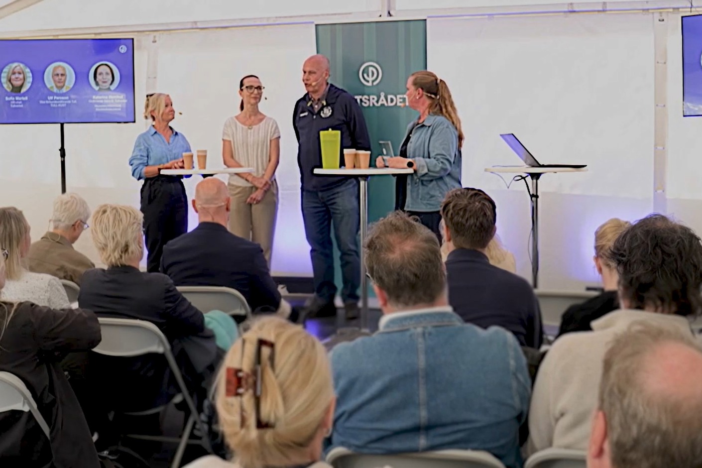 Tre personer står på en scen i ett tält och pratar med en fjärde som är moderator. Det är många i publiken.