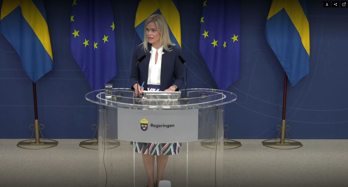 Person står vid ett glasbord som det står Regeringen på. Bakom syns två svenska och två EU-flaggor.